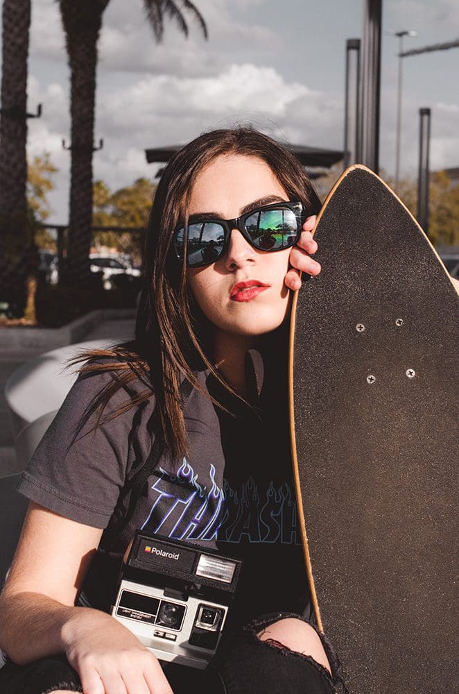 A lifestyle photo of a girl holding a camera and skateboard.