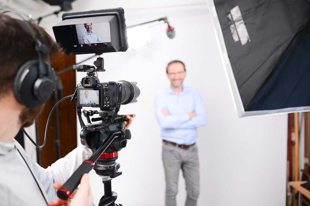 Camera operator filming a corporate interview subject in a studio with lights and monitor.
